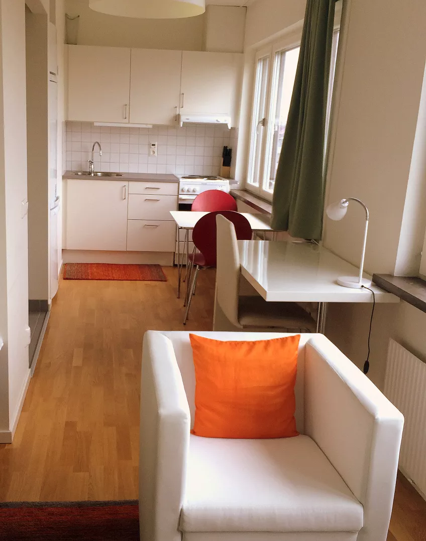 View of the kitchen from the living room in an apartment at the housing area Folkets park in the city of Malmö, with an armchair, a desk, and a kitchen table (picture).