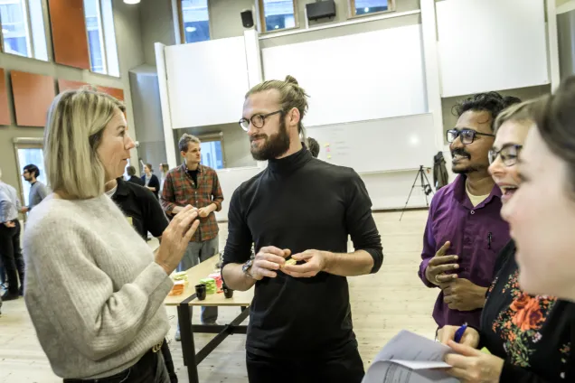 A woman talking to a man and three clustered people in a large space with whiteboards in the background. Photo by Kennet Ruona.