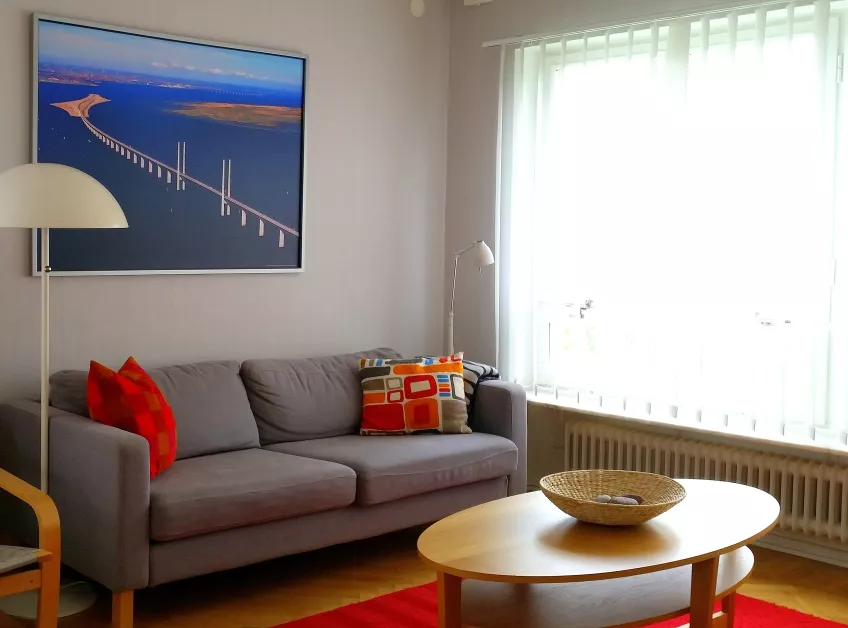 A picture of a living room at Gerdagatan 11 showing a sofa with a picture on the wall behind it, a coffee table, and a window (photo).