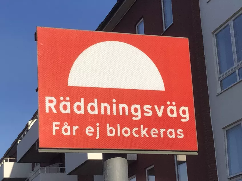 A red sign with white text saying "The escape route must not be blocked" in swedish". Behind it a building with several windows. (picture)