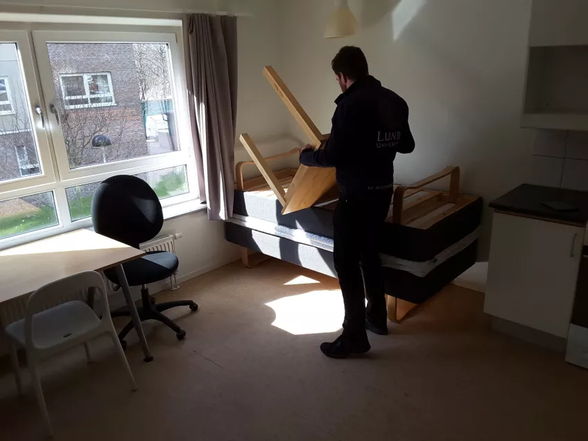 A picture showing an inspector turning a table upside down in a twin room at the housing area Klostergården.  Shown in the picture is also the two beds behind the inspector. One is turned upside down. The desk, and two chair are also visible to the left.