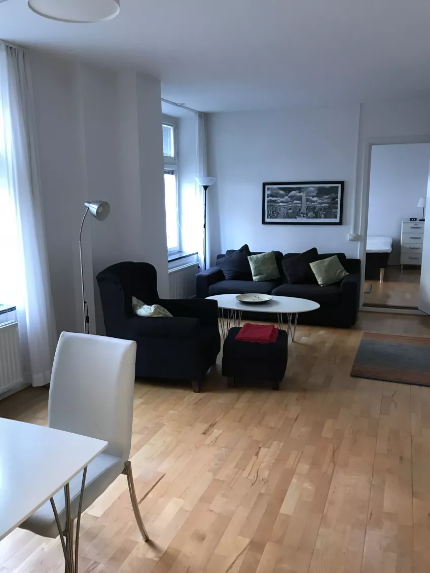 An overview of the living room at the housing area Kyrkogatan showing a chair to the left, armchair, sofa and coffee table in the middle and the door to the bedroom to the right (picture)