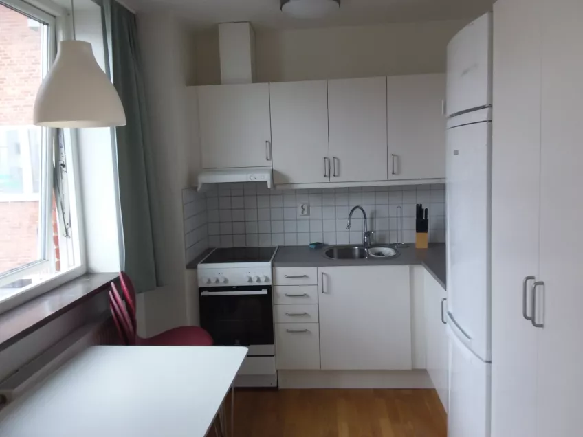 An overview of the kitchen in apartment 1210 at housing area Folkets Park showing the window and table to the left, stove and sink area in the middle and refrigerator and freezer to the right (picture)