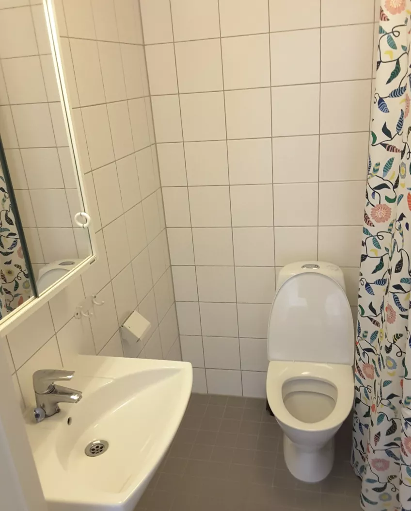 Overview of the bathroom in studio flat 1303 at housing area Folkets park showing the sink and mirror to the left, toilet in the middle and shower curtain to the right (picture)