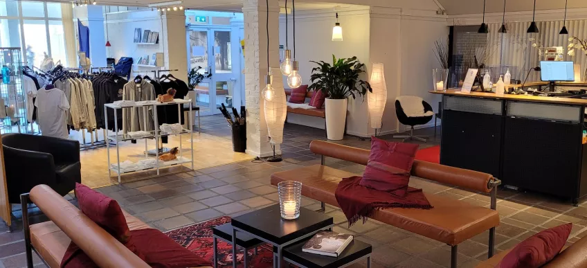 An overview of the reception area at University Guest House showing a lounge area with sofas and pillows in the middle, the Lund University Shop with clothes on racks to the left and the reception station to the right.
