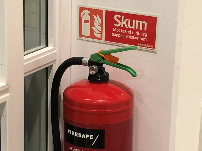 A red foam extinguisher below a sign showing where it is suppose to be hanging on a white wall next to two glass windows (picture)