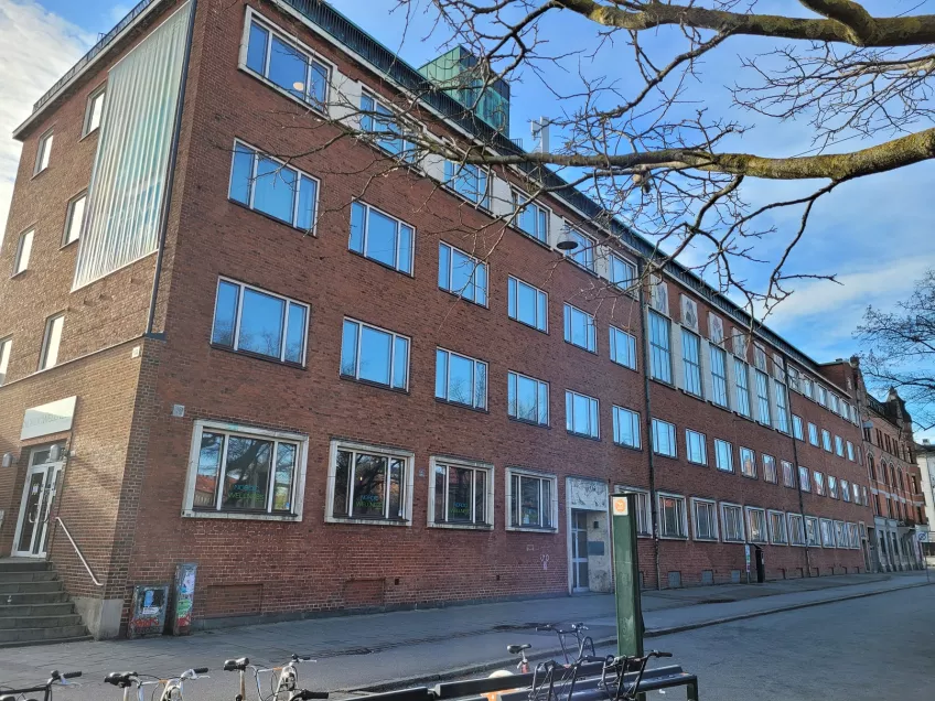 An overview of the housing area Folkets park in the city of Malmö showing the building where the apartments are. The building is built in reddish bricks, the windows reflect the blue sky on the top three floors (picture)