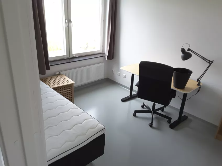 Overview of room 1407 B at the housing area Sofieberg showing the private room with the bed to the left and window beyond it, desk chair, desk and desk lamp to the right (picture).