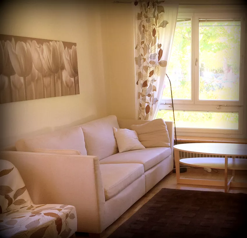 A picture of a living room at Gerdagatan 13 showing a sofa with a picture on the wall behind it, a coffee table, and a window with curtains (photo).