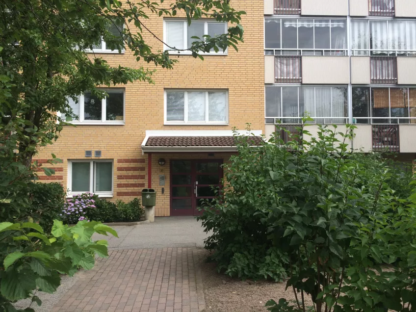 The entrance to Luzernvägen 10 seen from the front, a three storey building with trees and bushes in front (photo).