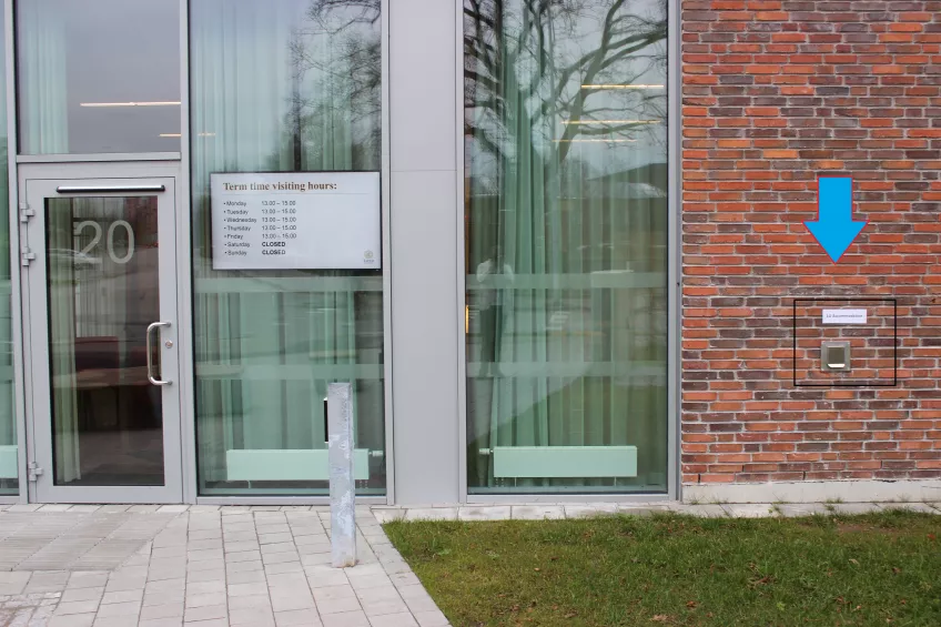 A glass reception entrance in a red brick building, with an added blue arrow pointing out a key return chute to the right of the entrance. Edited photo by LU Accommodation.